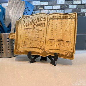 Puede incluir: Una tabla de cortar de madera con forma de libro con temática de bruja. La tabla incluye una tabla de conversión para tazas, onzas, cucharadas, cucharaditas y mililitros. También incluye una tabla de conversión de temperatura y peso. La tabla está decorada con un caldero de bruja, botellas y una telaraña.