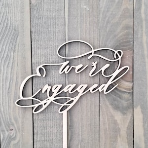 Op de afbeelding: Een houten taarttopper met de woorden "we're engaged" in een sierlijk lettertype. De topper is licht van kleur en heeft een dunne stok om in een taart te steken. De achtergrond is een grijze houten ondergrond.