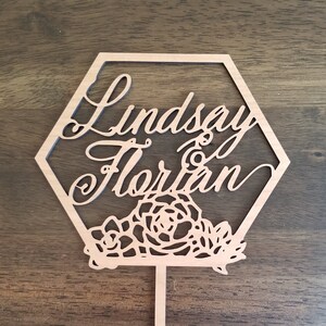 Op de afbeelding: Een houten zeshoekige taarttopper met de namen "Lindsay & Florian" in cursief schrift. De topper heeft een bloemmotief met ingewikkelde uitsnijdingen.