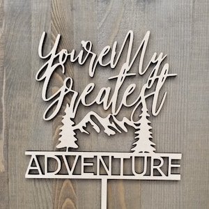 Op de afbeelding: Houten taarttopper met de tekst "You're My Greatest Adventure" en een silhouet van bergen en bomen.