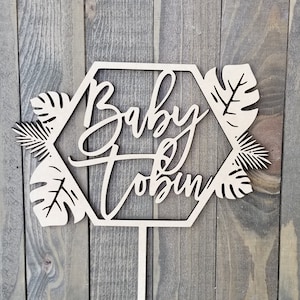 Op de afbeelding: Houten taarttopper met een zeshoekige vorm en tropische bladeren. De zeshoek bevat de tekst "Baby Tobin".
