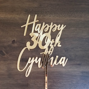Op de afbeelding: Gouden glitter taarttopper met de tekst "Happy 30th Cynthia".