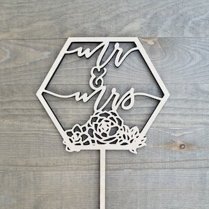Op de afbeelding: Een houten taarttopper in de vorm van een zeshoek met de woorden "Mr & Mrs" geschreven in een cursief schrift. De topper is versierd met een bloemmotief en heeft een houten stok om in een taart te steken.
