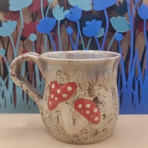 May include: A ceramic mug with a speckled cream and brown glaze featuring two raised red mushrooms with white spots on the front. The mug has a curved handle and a wide rim with a smooth, glossy finish that drips down towards the textured base.