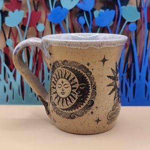 May include: A speckled tan ceramic mug featuring a black ink illustration of a sun and moon face surrounded by swirling clouds and small stars. The mug has a sturdy handle and a light-coloured glazed rim, showcasing a detailed celestial design.
