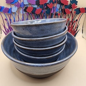 May include: A stack of four ceramic bowls with a speckled blue and grey glaze. The bowls feature a smooth, rounded rim and a deep basin. Behind the bowls, a decorative arrangement of thin, upright metal stems topped with colourful flower shapes.