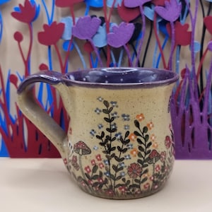 May include: A ceramic mug with a speckled beige base and a dark purple interior rim. The exterior features a hand-painted botanical design with small red, orange, and blue flowers, green stems, and two small red-capped mushrooms near the base.