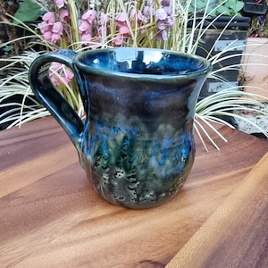 May include: A ceramic mug featuring a glossy glaze with a blend of deep blue, forest green, and black tones. The mug has a rounded body, a wide rim, and a curved handle on the side. The surface texture shows variations in colour intensity and depth.