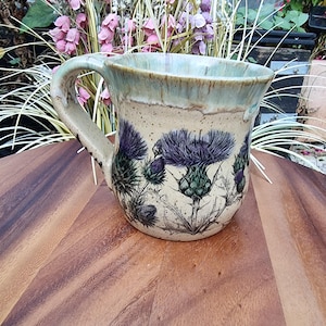 May include: A ceramic mug with a speckled cream-coloured base featuring detailed illustrations of purple thistle flowers. The mug has a curved handle and a wide rim with a light blue-green glaze that drips down the upper portion of the exterior.
