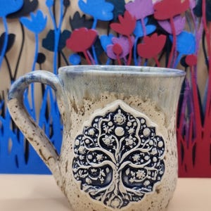 May include: A ceramic mug with a speckled cream and blue glaze featuring a raised, dark blue circular medallion on the front. The medallion displays a detailed white relief of a tree with intricate branches, leaves, and small circular floral motifs.