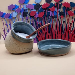 May include: A ceramic salt cellar with a speckled beige exterior and a smooth blue interior, containing a wooden spoon. Beside it sits a matching blue ceramic spoon rest. The set is presented against a backdrop of colourful felt flowers.