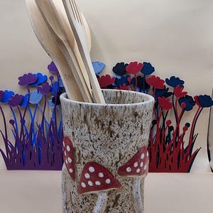May include: A cylindrical ceramic utensil holder with a speckled beige finish, featuring raised red mushroom designs with white spots. Several light-coloured wooden kitchen utensils are placed inside the holder, which stands in front of decorative cutouts of red and blue flowers.