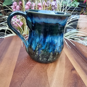 May include: A ceramic mug featuring a glossy glaze with a deep blue and dark green colour palette. The surface displays a drip effect pattern that transitions from a lighter blue at the rim to a darker, forest-like green at the base of the vessel.