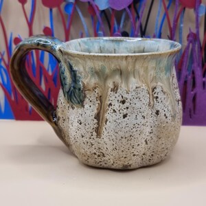 May include: A ceramic mug with a rounded, pumpkin-like shape featuring a speckled cream and tan base. The upper rim has a glossy, drippy glaze in shades of blue and brown, with a matching dark brown curved handle and a decorative leaf accent.