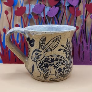 May include: A speckled tan ceramic mug featuring a black line art illustration of a rabbit decorated with floral patterns. The mug has a wide handle and a white interior rim. The rabbit design includes small flowers and leaves within its body.