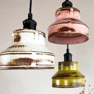 May include: Three pendant lights with a rustic, vintage look. The lights are made of metal and have a distressed finish. The shades are white, pink, and yellow.