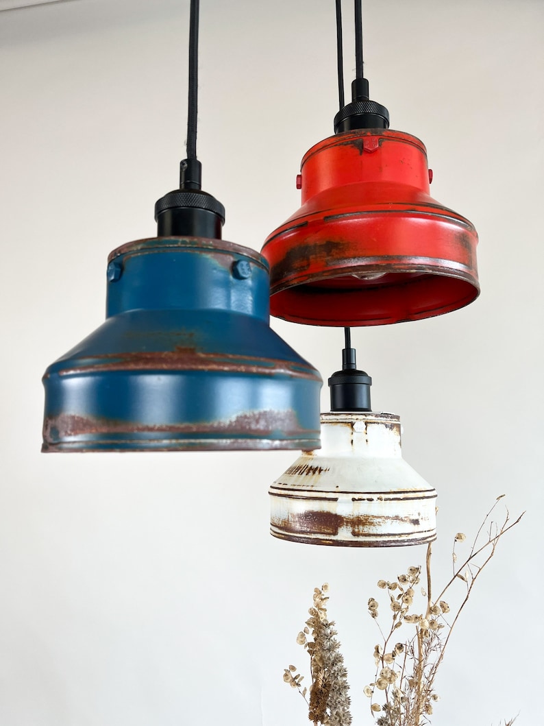 May include: Three pendant lights with a rustic, industrial style. The lights are made of metal and have a distressed finish. The shades are red, blue, and white.