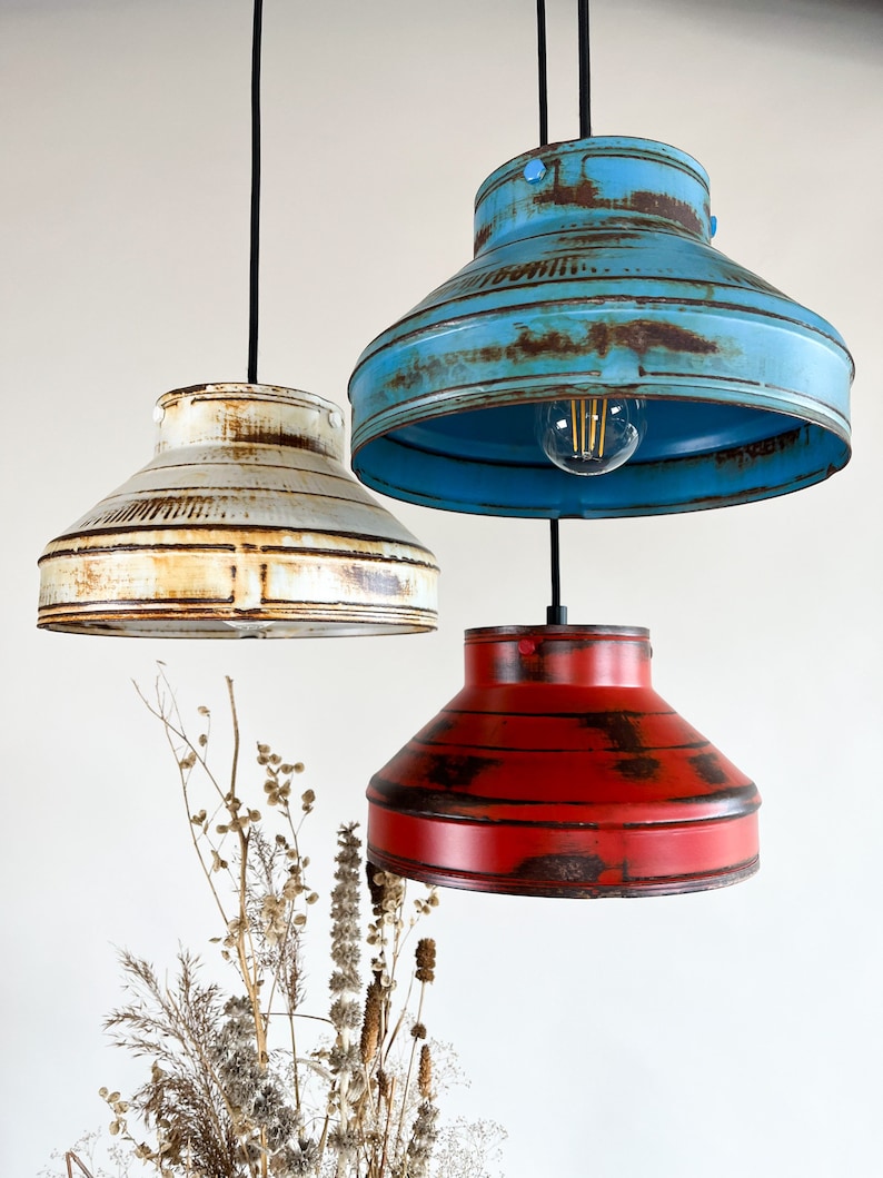 May include: Three pendant lights with distressed metal shades in blue, white, and red. The shades are round and have a slight curve to them. The lights are hanging from black cords.