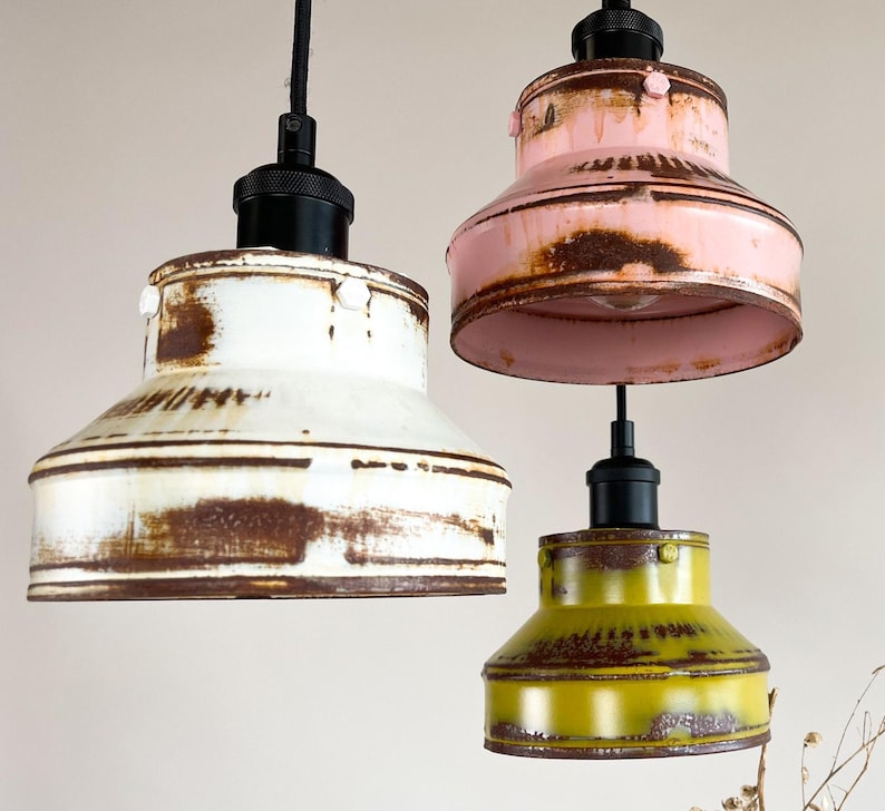 May include: Three pendant lights with a rustic, vintage look. The lights are made of metal and have a distressed finish. The shades are white, pink, and yellow.