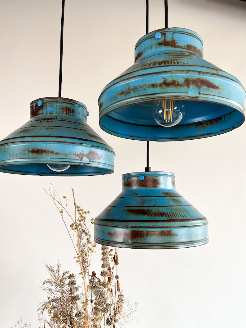 May include: Three blue metal pendant lights with a distressed finish. The lights have a wide, rounded shape and are hanging from black cords.
