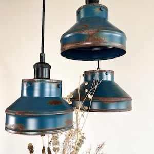 May include: Three blue pendant lights with a distressed, rusty finish. The lights are hanging from black cords and are suspended over a bouquet of dried flowers.