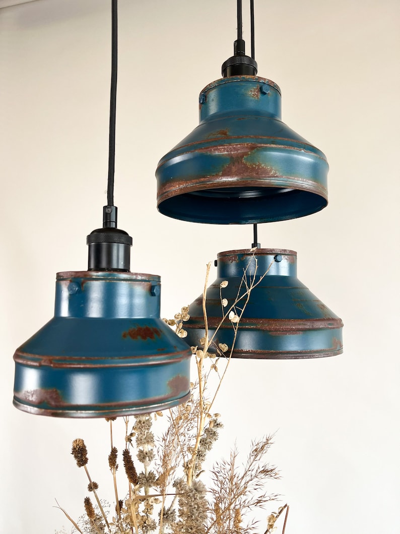 May include: Three blue pendant lights with a distressed, rusty finish. The lights are hanging from black cords and are suspended over a bouquet of dried flowers.