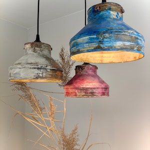 May include: Three hanging pendant lights with distressed metal shades in blue, white, and pink. The shades have a rustic, aged appearance. The lights are suspended from black cords against a neutral wall, with dried ornamental grass in the foreground.