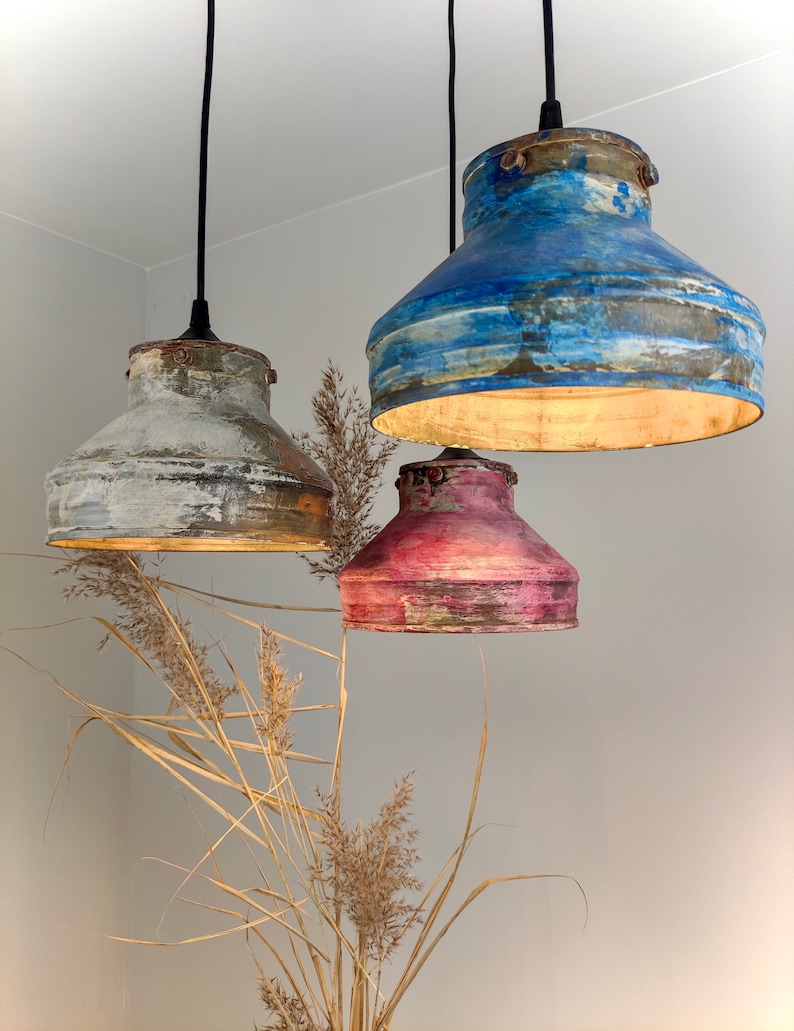 May include: Three hanging pendant lights with distressed metal shades in blue, white, and pink. The shades have a rustic, aged appearance. The lights are suspended from black cords against a neutral wall, with dried ornamental grass in the foreground.