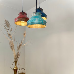 May include: Three pendant lights with distressed metal shades in red, turquoise, and blue. Each light hangs from a black cord and illuminates with a warm glow. The lights are suspended in a corner of a room, with a vase and decorative items in the foreground.