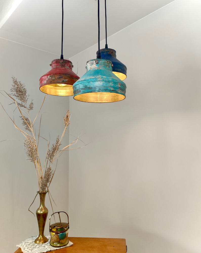 May include: Three pendant lights with distressed metal shades in red, turquoise, and blue. Each light hangs from a black cord and illuminates with a warm glow. The lights are suspended in a corner of a room, with a vase and decorative items in the foreground.
