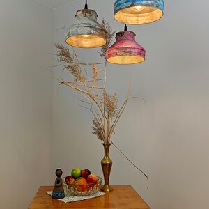 May include: Three hanging pendant lights with distressed paint finishes in blue, pink, and white. The lights are suspended above a wooden table with a vase of dried flowers and a bowl of fruit. A decorative figurine is on the table.