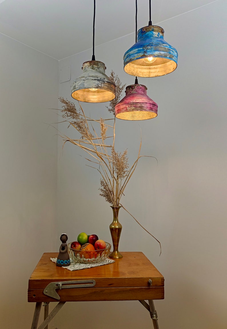 May include: Three hanging pendant lights with distressed paint finishes in blue, pink, and white. The lights are suspended above a wooden table with a vase of dried flowers and a bowl of fruit. A decorative figurine is on the table.