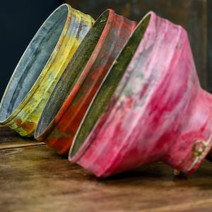 May include: Close-up of three vintage-style metal lamp shades in yellow, red, and pink. The shades are arranged diagonally, showcasing their aged, distressed appearance. The brass lamp holder is visible on the right, suggesting a focus on industrial or rustic lighting.