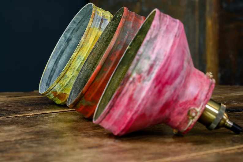 May include: Close-up of three vintage-style metal lamp shades in yellow, red, and pink. The shades are arranged diagonally, showcasing their aged, distressed appearance. The brass lamp holder is visible on the right, suggesting a focus on industrial or rustic lighting.