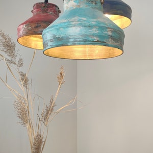 May include: Three hanging pendant lights with distressed metal shades in red, teal, and navy blue. The interior of each shade is gold, and the lights are suspended from black cords. A vase of dried plants is in the foreground.