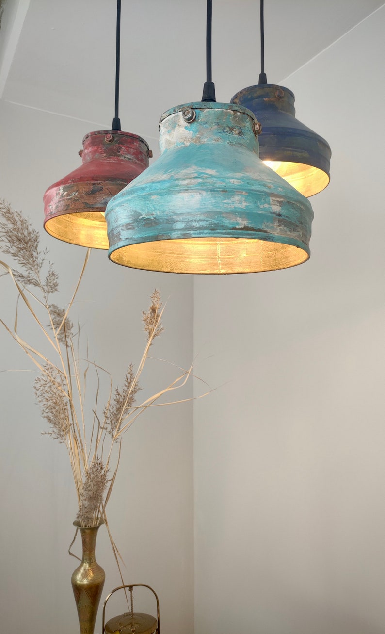 May include: Three hanging pendant lights with distressed metal shades in red, teal, and navy blue. The interior of each shade is gold, and the lights are suspended from black cords. A vase of dried plants is in the foreground.
