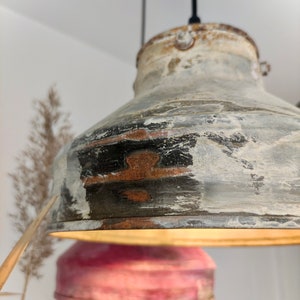 May include: A distressed metal pendant light with a rustic, industrial aesthetic. The light fixture features a weathered, multi-toned finish with shades of gray, black, and white, and a hint of rust. The light is suspended from a black cord.