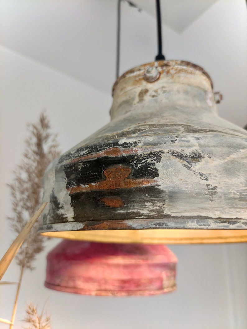 May include: A distressed metal pendant light with a rustic, industrial aesthetic. The light fixture features a weathered, multi-toned finish with shades of gray, black, and white, and a hint of rust. The light is suspended from a black cord.