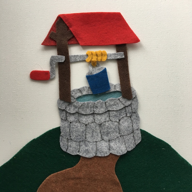 May include: A felt well with a red roof, gray stone walls, and a blue bucket. The well is set against a white background with a green hill and brown base. The well has a wooden frame and a yellow rope.