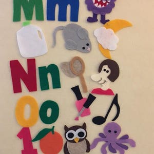 May include: Felt letter set for children, featuring the letters M, N, O, and a 1. The set includes various felt images, including a monster, a mouse, a milk carton, a net, a woman, a nail polish bottle, a musical note, an owl, an orange, and an octopus.