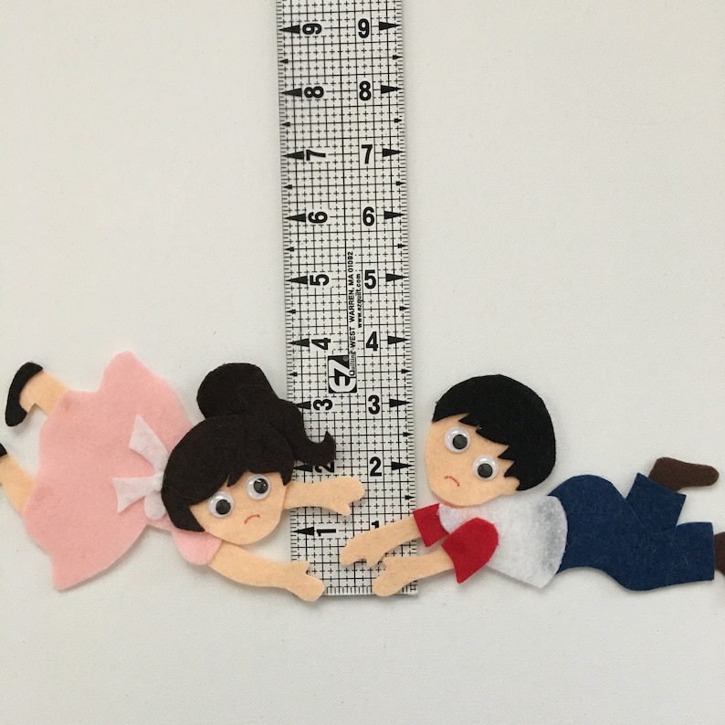 May include: Felt figures of a girl in a pink dress and a boy in a white shirt and blue pants, both with sad expressions. A ruler shows the figures are approximately 3 inches in length.