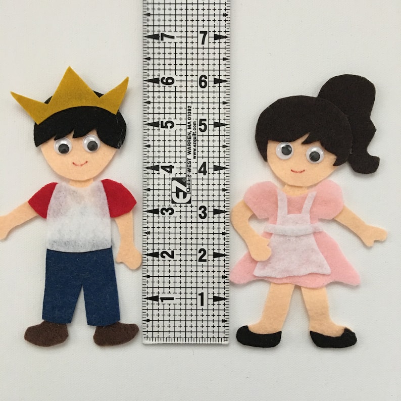 May include: Felt figures of a boy and girl, with a ruler for scale. The boy wears a gold crown, red and white shirt, and blue pants. The girl is in a pink dress and apron. The ruler shows the figures are approximately 3 inches tall.