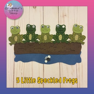 May include: A felt craft depicting five green speckled frogs sitting on a brown log above blue water. The text "5 Little Speckled Frogs" is at the bottom. The image has a pink and purple gradient background.