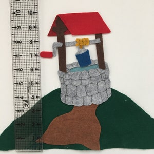 May include: A felt well with a red roof, brown posts, and a gray stone base. A blue bucket hangs from a yellow rope. The well is on a green hill, with a brown tree trunk. The ruler shows the well is approximately 7 inches tall.