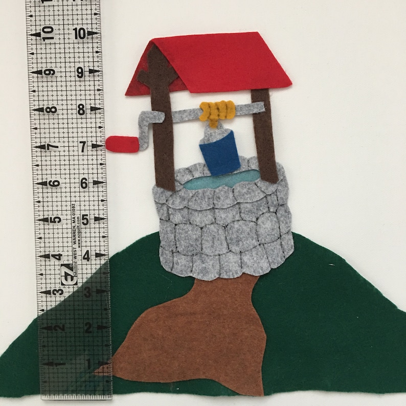 May include: A felt well with a red roof, brown posts, and a gray stone base. A blue bucket hangs from a yellow rope. The well is on a green hill, with a brown tree trunk. The ruler shows the well is approximately 7 inches tall.
