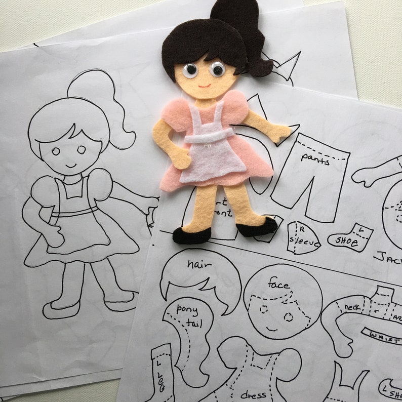 May include: A felt doll with a pink dress and white apron, black shoes, and a brown ponytail, sits atop paper templates for a paper doll. The templates include outlines for clothing and body parts, labeled with terms like "pants", "sleeve", and "dress".