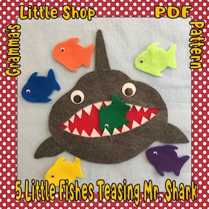 May include: A felt craft pattern for a shark with an open mouth and five little fish. The shark is gray with white teeth and big eyes. The fish are orange, blue, green, yellow, and purple. The pattern is titled "5 Little Fishes Teasing Mr. Shark" and "Grandma's Little Shop PDF pattern".