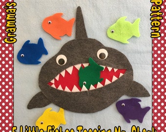 May include: A felt board featuring a gray shark with a red mouth and white teeth, surrounded by five colorful fish. The text "5 Little Fishes Teasing Mr. Shark" is displayed at the bottom. The words "Little Shop" and "PDF Pattern" are at the top.