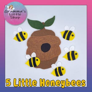 May include: A felt craft depicting a brown beehive with green leaves and five yellow and black bees. The text "5 Little Honeybees" is at the bottom. The image has a pink and purple gradient border and a logo in the upper left corner.