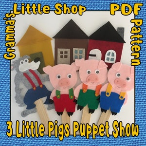 May include: A felt puppet show set featuring three little pigs, a wolf, and three houses. The houses are yellow, brown, and red. The puppets are attached to wooden sticks. The text "Grandma's Little Shop PDF Pattern 3 Little Pigs Puppet Show" is printed on the image.